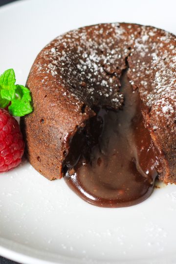cut open chocolate lava cake with powdered sugar on top and raspberries and mint beside it.
