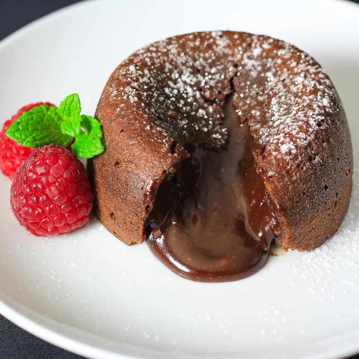 cut open chocolate lava cake with powdered sugar on top and raspberries and mint beside it.