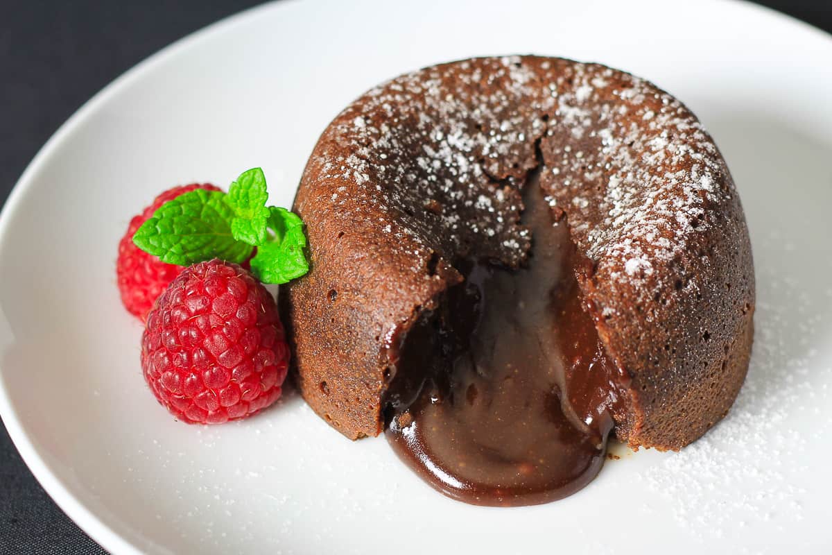 cut open chocolate lava cake with powdered sugar on top and raspberries and mint beside it.