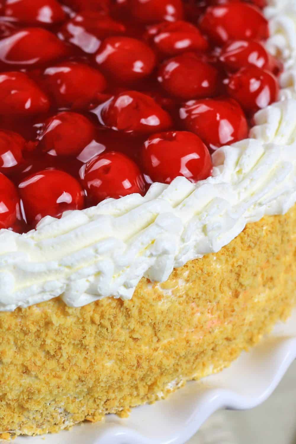 Up close picture of a completed cherry jello cake recipe with the cake sitting on a white cake plate and topped with cherry pie filling, cream cheese filling, and a coating of cake crumbs around the edges