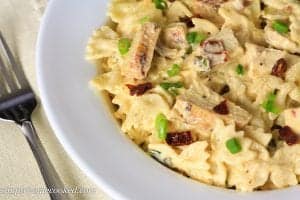 A white bowl of alfredo with chicken, green onions, and sun dried tomatoes. A fork next to the bowl.