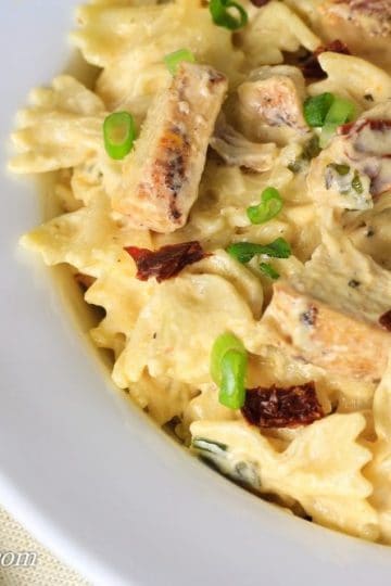 A white bowl of alfredo with chicken, green onions, and sun dried tomatoes. A fork next to the bowl.