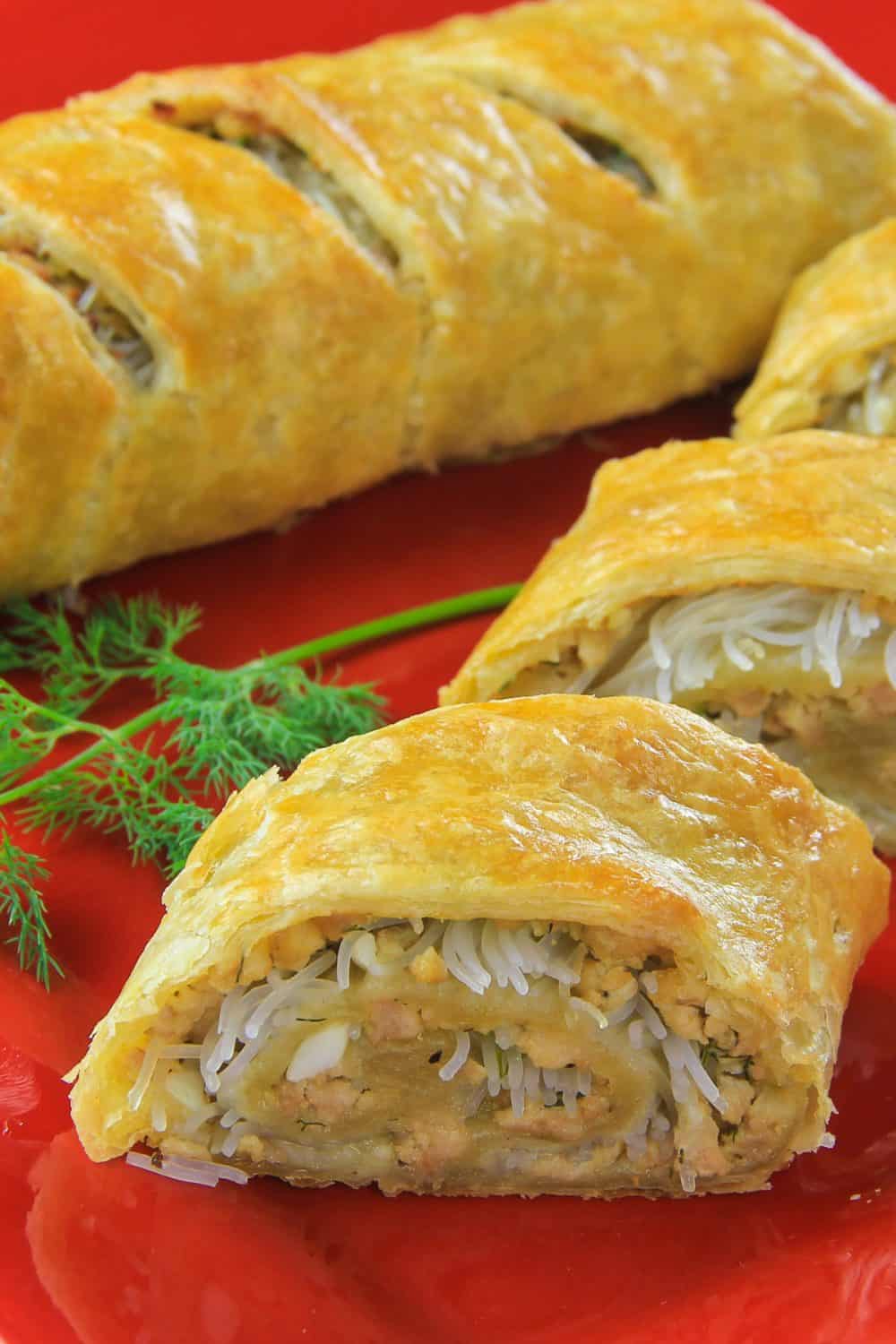 Prepared chicken puff pastry stuffed with rice noodles, eggs, dill, and ground chicken presented on a red plate with fresh dill as a garnish