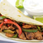 chicken fajitas on a white plate with a bowl of sour cream and to limes next to it