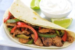 chicken fajitas on a white plate with a bowl of sour cream and to limes next to it