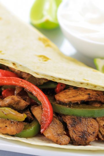chicken fajitas on a white plate with a bowl of sour cream and to limes next to it