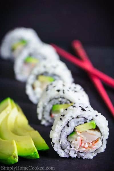 sliced shrimp tempura sushi roll with sliced avocado on the side along with red chopsticks