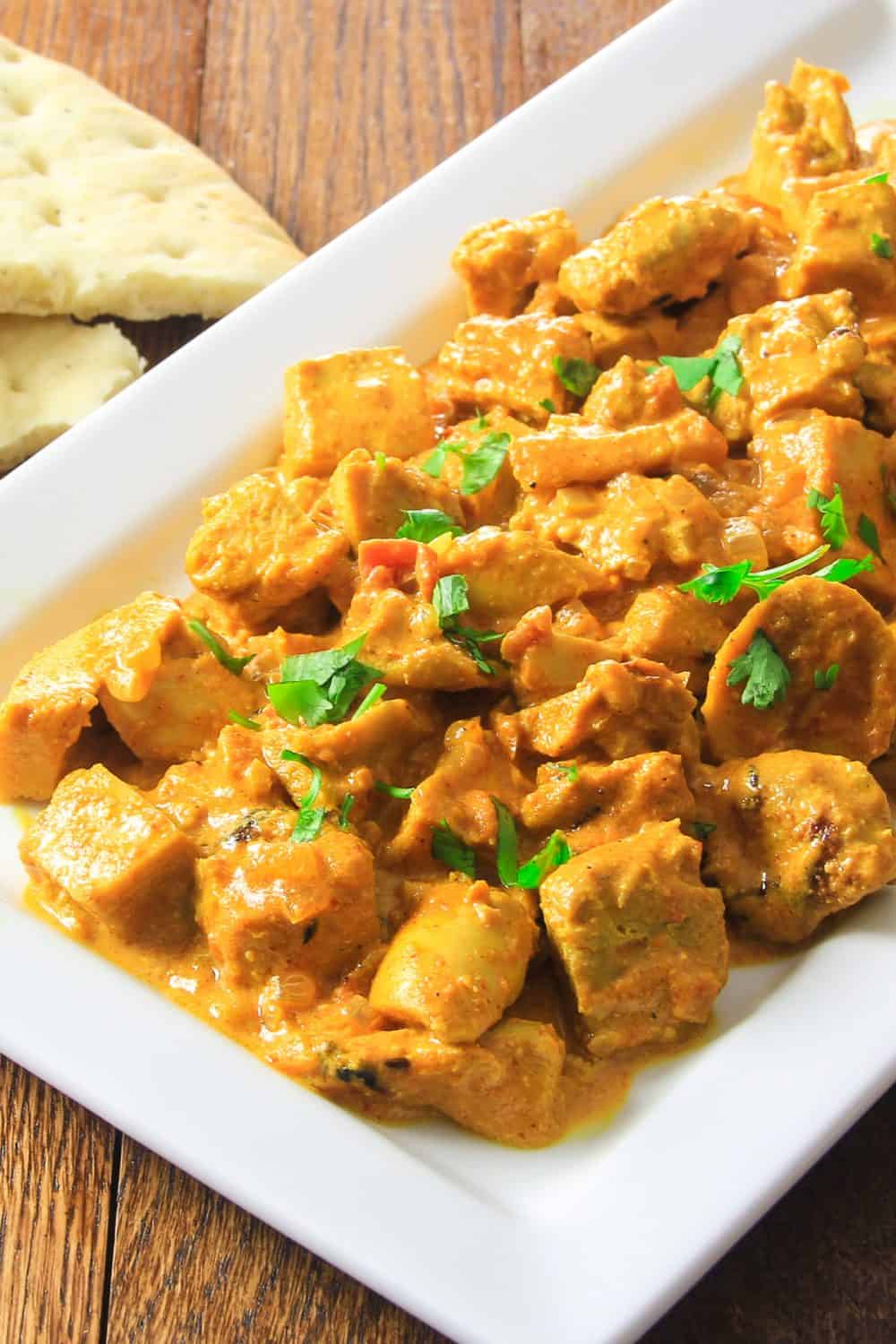 Close up of white plate filled with Indian Butter chicken with some parsley on top and bread to the side.