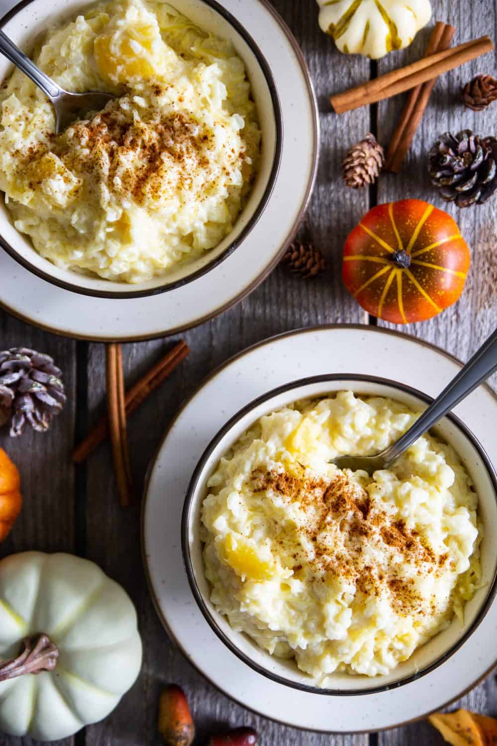 an overhead image of two bowls of rice pudding with cinnamon sticks and mini pumpkin beside them on a dark wooden background 
