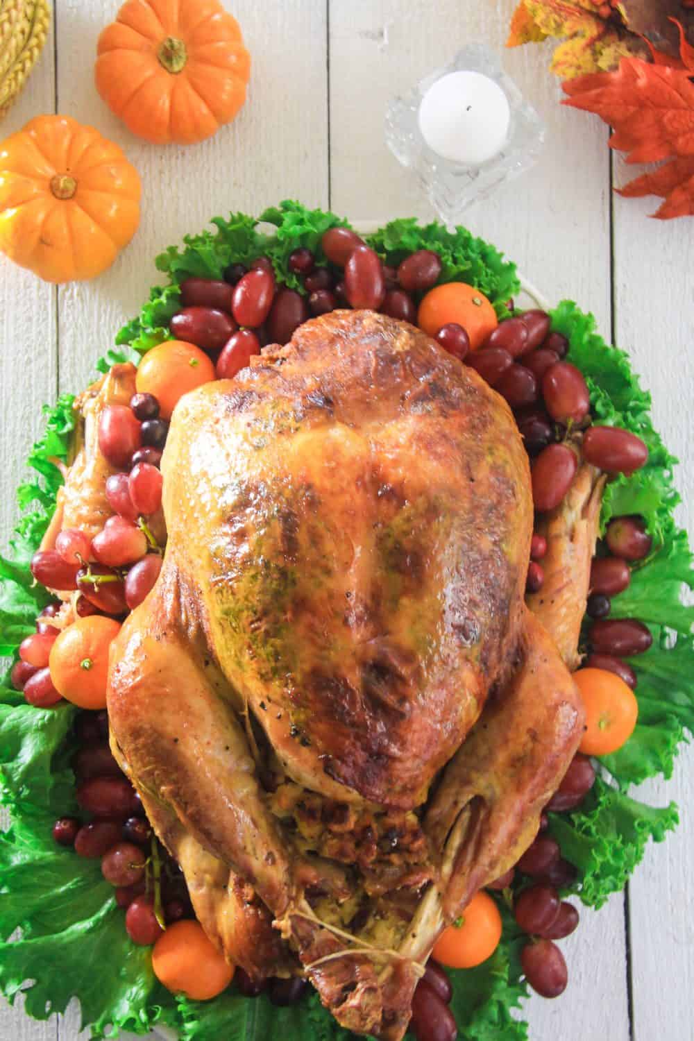 an overhead image of a whole roasted turkey on a white wooden background with grapes, pumpkins and Mandarins around it