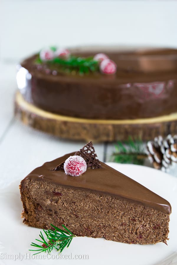 Close up slice of chocolate cherry cake on a white plate topped with a sugared cherry and chocolate slice with full cake in background on piece of wood laying on a white surface. 