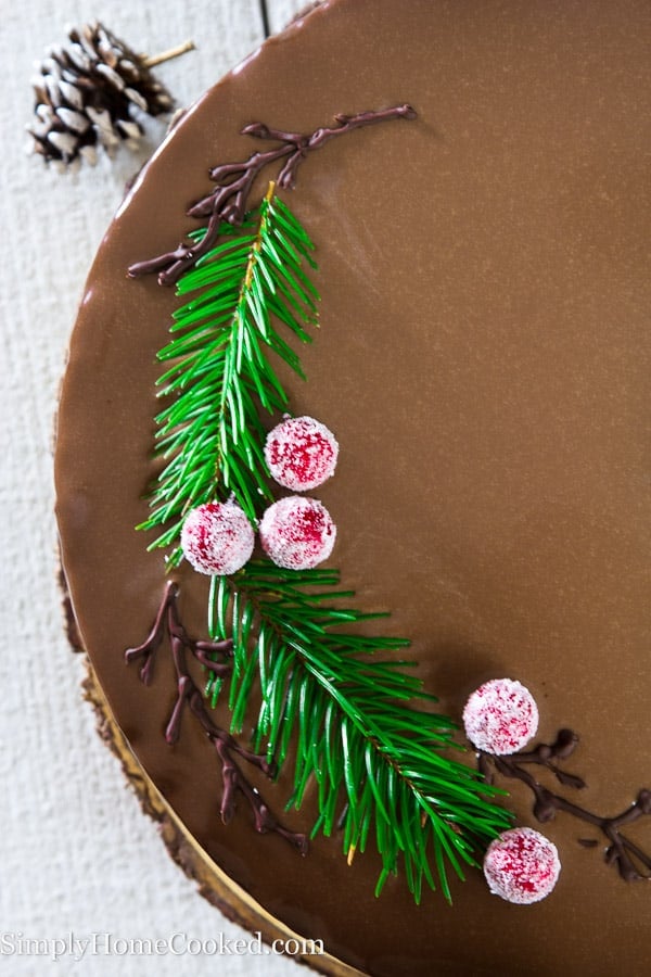 Overhead picture showing half of drunken chocolate cherry cake topped with greenery, sugared cherries, and piped chocolate branches while sitting on a white wooden surface. 
