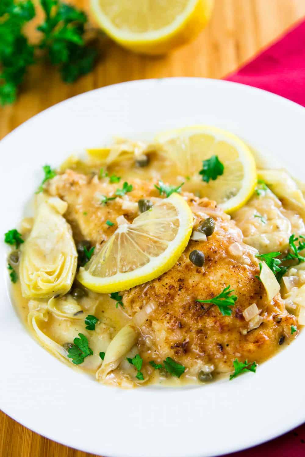 Lemon chicken piccata with a lemon slice on top in a white dish with a lemon and some Italian herbs in the background