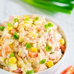potato salad with baked salmon in a white bowl with a slice of tomato and a bunch of green onion next to it.