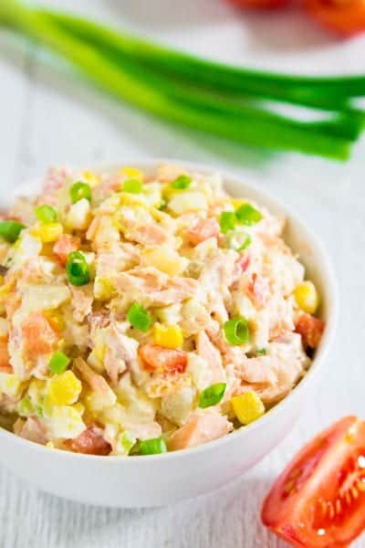 potato salad with baked salmon in a white bowl with a slice of tomato and a bunch of green onion next to it.