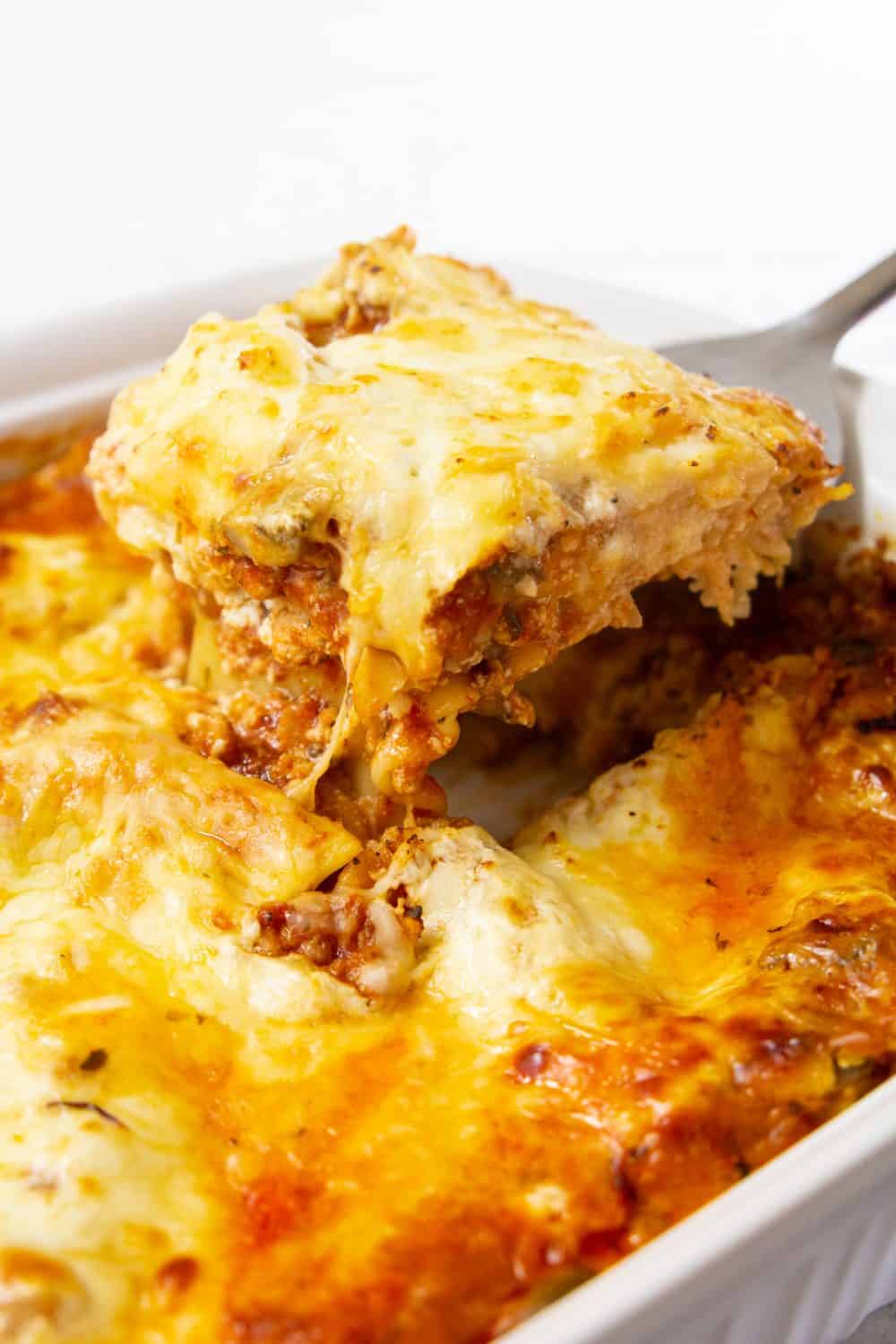 Close up image of cooked chicken lasagna recipe with one portion being scooped out with a spatula showing the melted cheese and layers of pasta and meat sauce