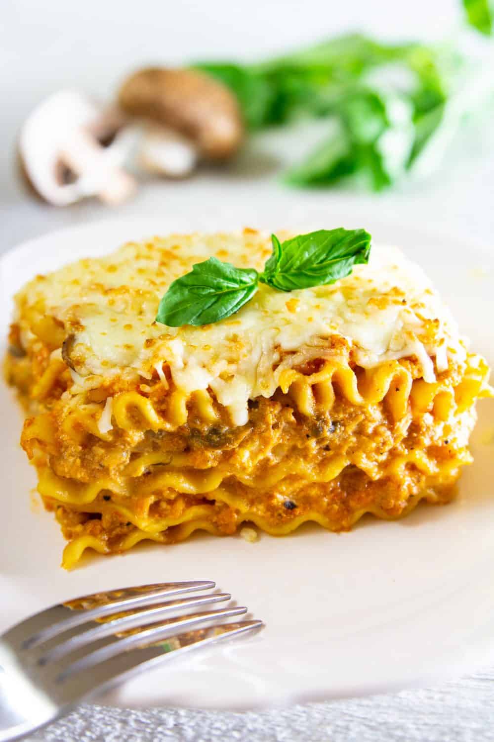 Chicken lasagna recipe serving on a white plate laying on a white surface with fork laying on the plate and a sprig of basil on top of lasagna