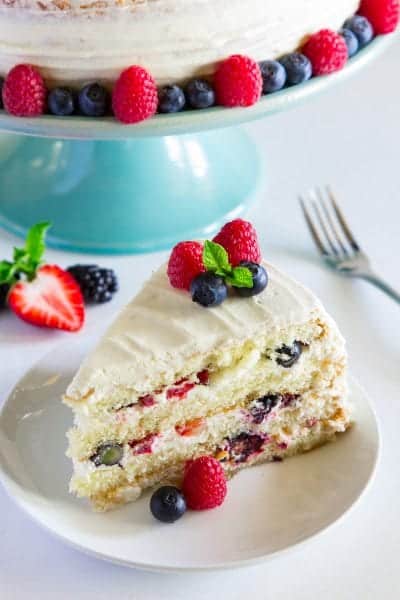 sliced Chantilly cake with berries on top on a white plate with a blue cake stand behind it