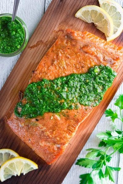 grilled salmon on cedar plank with chimichuri sauce on top and sliced lemons beside it.