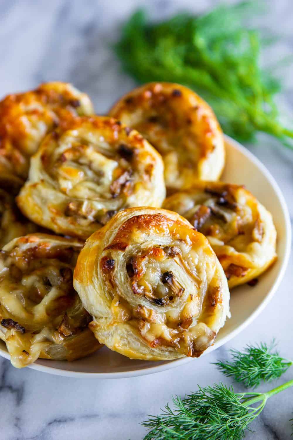 Up close image of Mushroom Puff Pastry Pinwheels on a white platter sitting on a marble counter with fresh herbs beside the platter. 