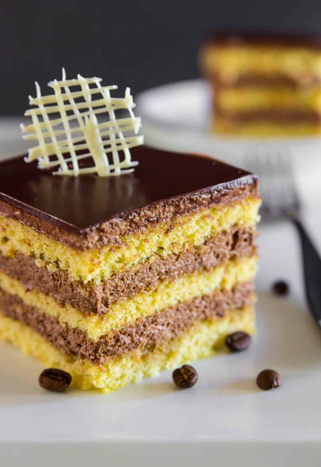 a close up image of a homemade opera cake sliced into a square with coffee beans beside it and a fork behind it