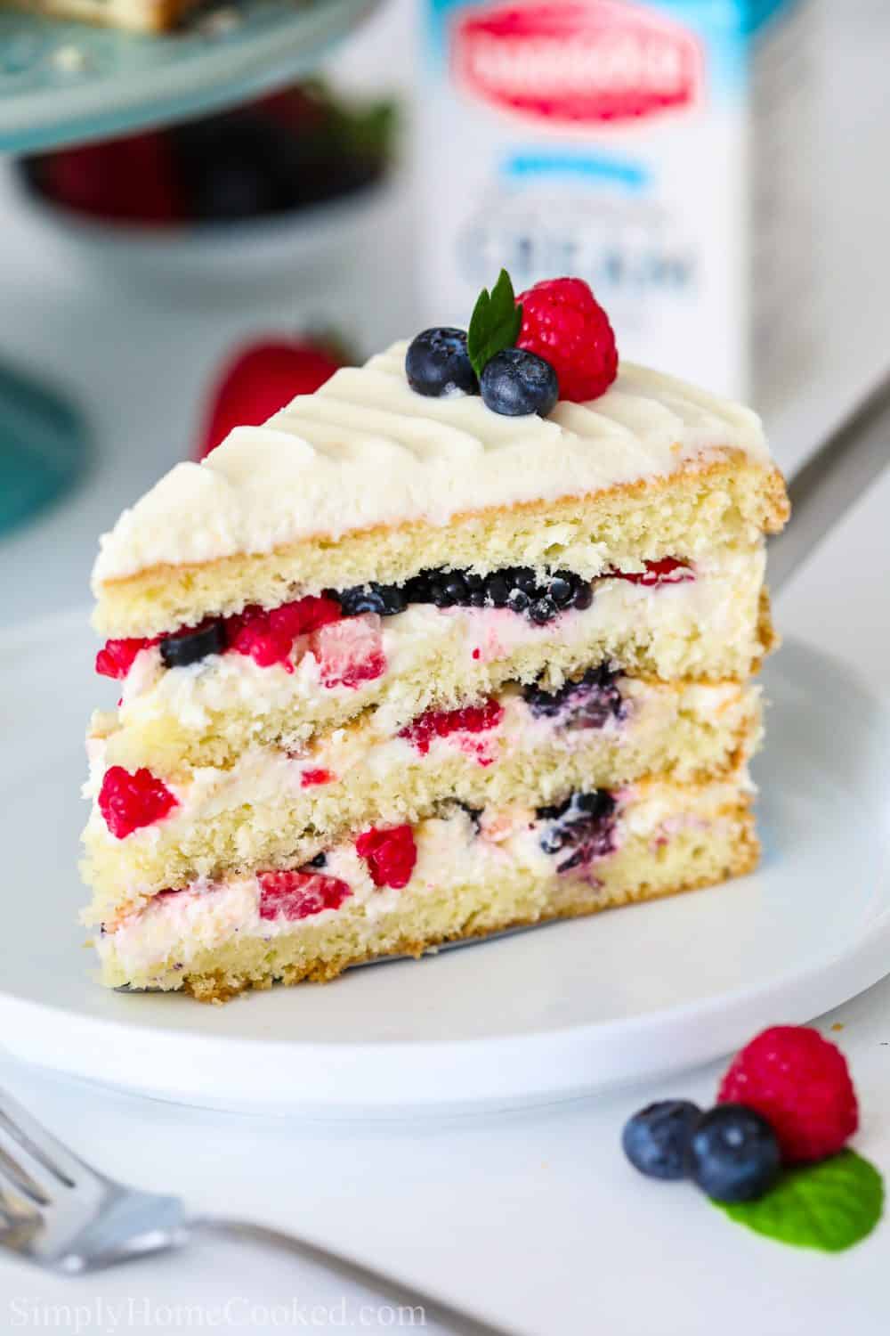 sliced Chantilly cake with berries on top on a white plate with a blue cake stand behind it