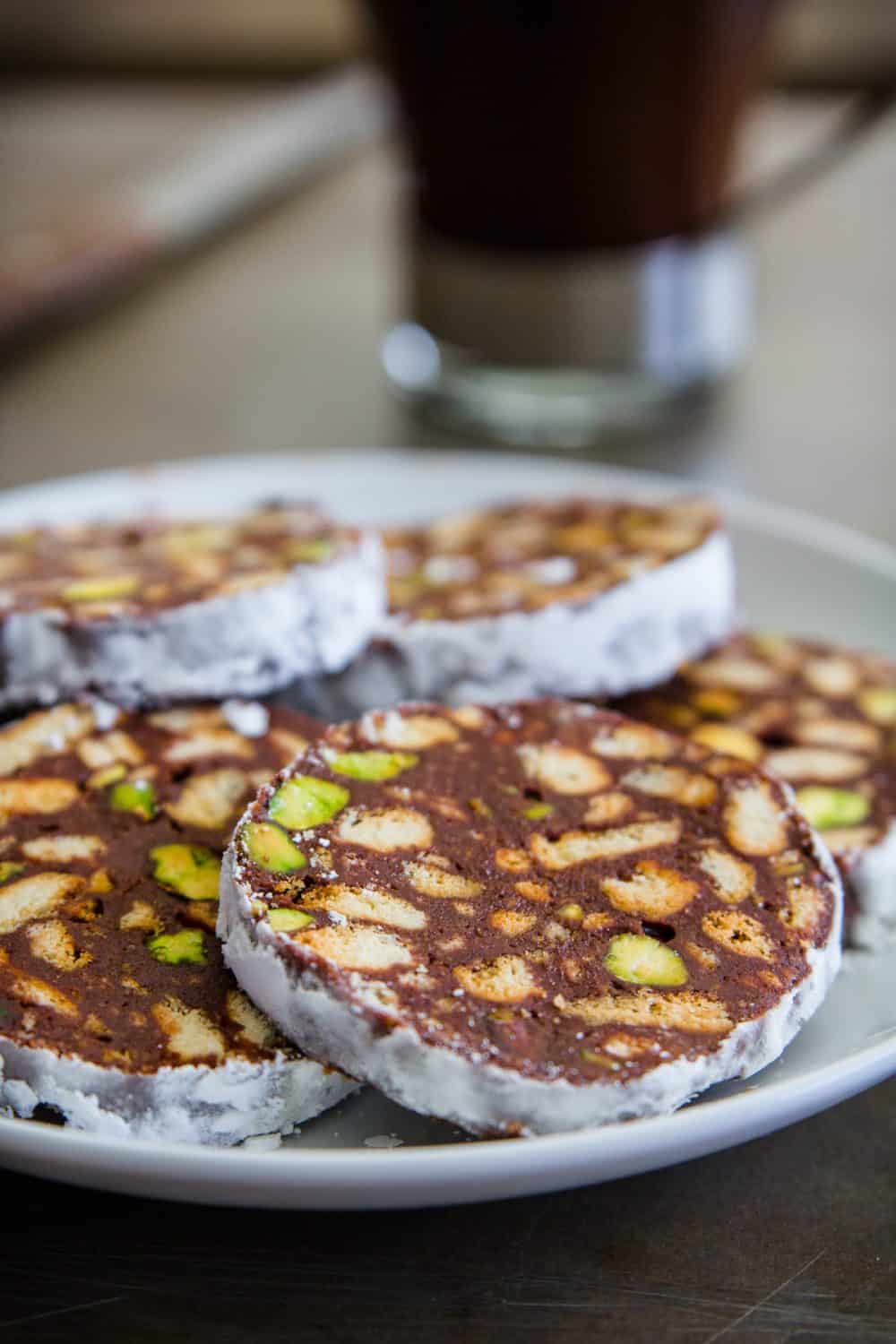 sliced chocolate, biscuit, and pistachios salami on a white plate with a cup of coffee behind it and a knife beside it