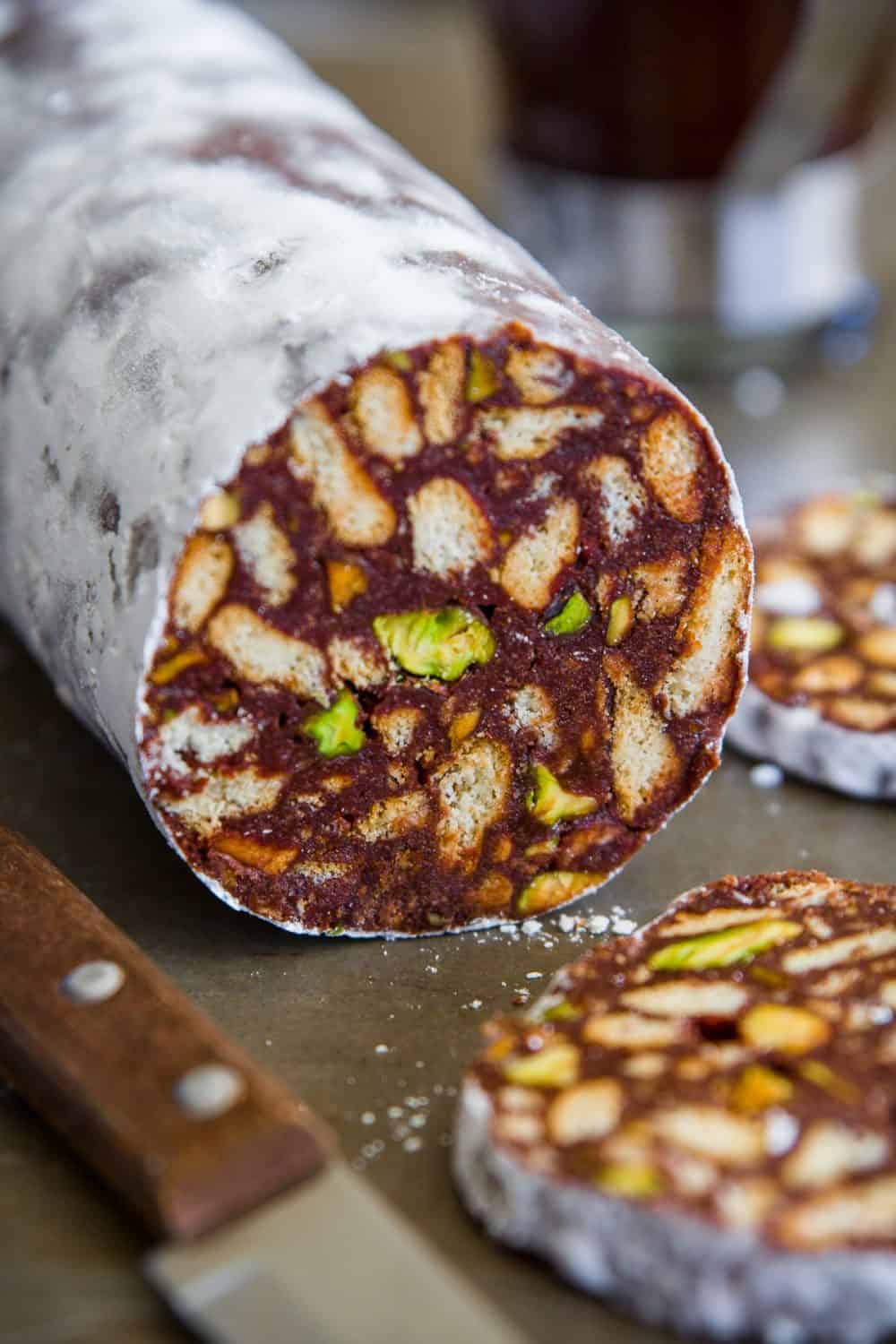 sliced chocolate, biscuit, and pistachios salami on a white plate with a cup of coffee behind it and a knife beside it