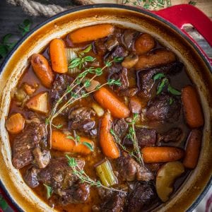 braised beef stew in a red dutch oven pot with herbs beside it.