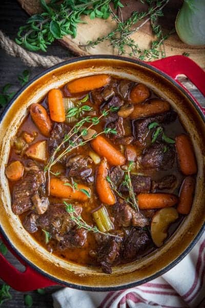 braised beef stew in a red dutch oven pot with herbs beside it.
