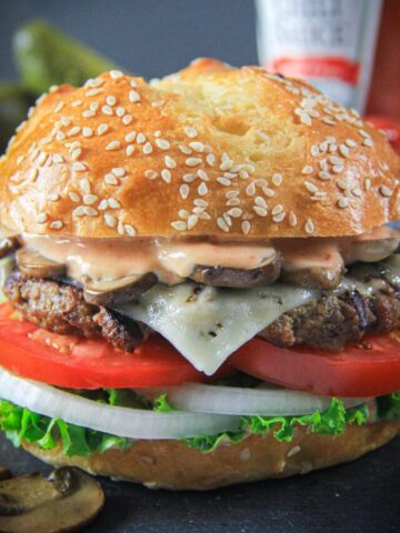 Burger Patty recipe on a bun with lettuce, tomato, cheese, onion, mushrooms, and sauce with ketchup in the background.