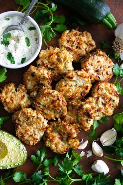 paleo chicken zucchini fritters with garlic cloves , sliced avocado, and parsley beside it.