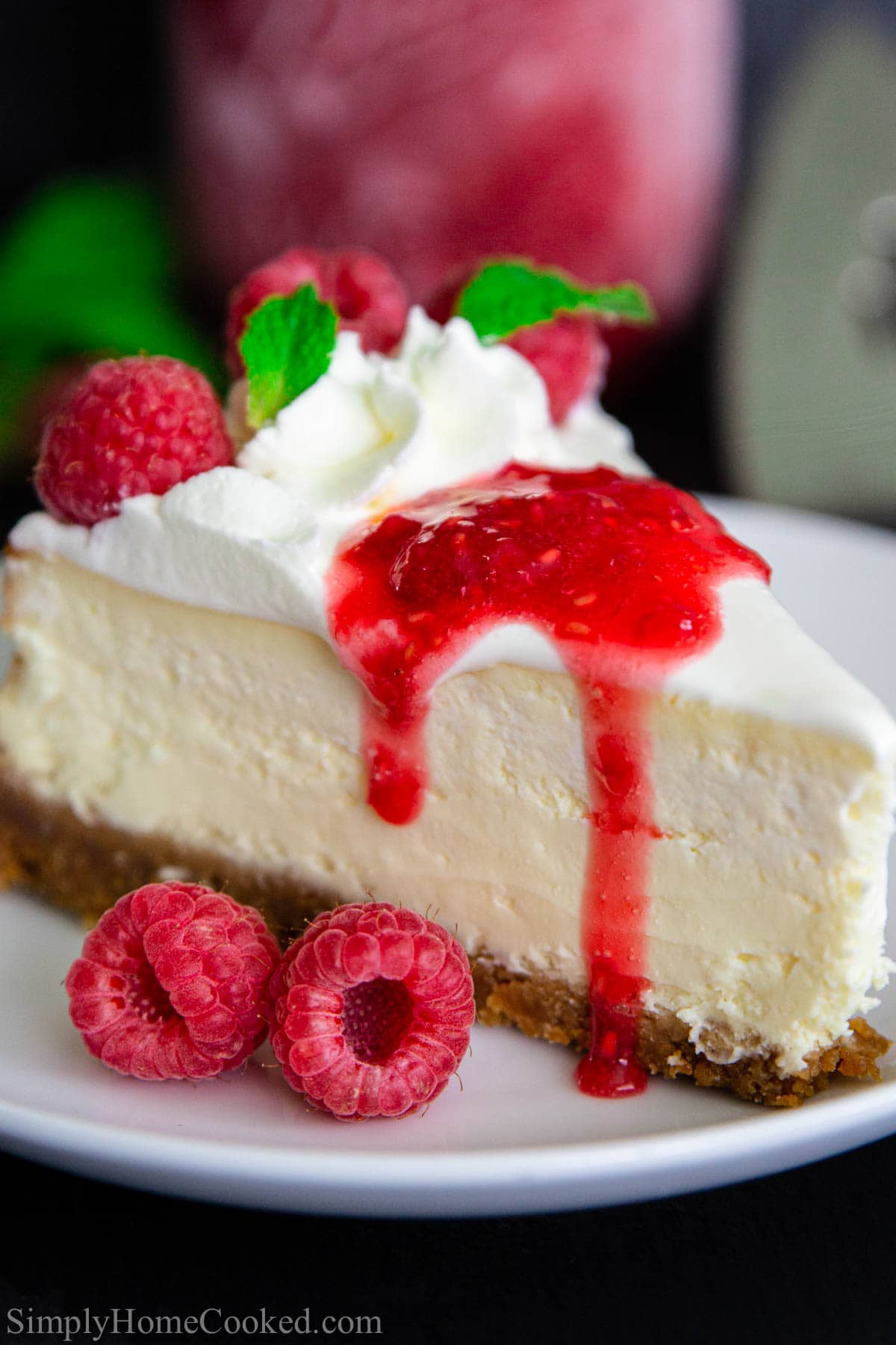 A slice of New York style cheesecake on a white plate with raspberry sauce dripping over.