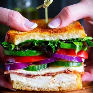 two hands holding a sandwich with lettuce, tomatoes, red onion, bacon, and sour dough bread