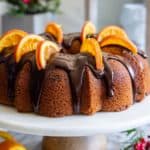 cranberry orange bundt cake with chocolate ganache on top, and dried orange slices on top, with an orange peal beside it