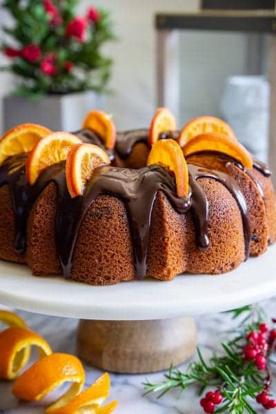 cranberry orange bundt cake with chocolate ganache on top, and dried orange slices on top, with an orange peal beside it