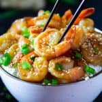 garlic ginger shrimp with green onion and sesame seeds on top in a white bowl with black chop sticks picking up the shrimp