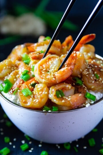 garlic ginger shrimp with green onion and sesame seeds on top in a white bowl with black chop sticks picking up the shrimp