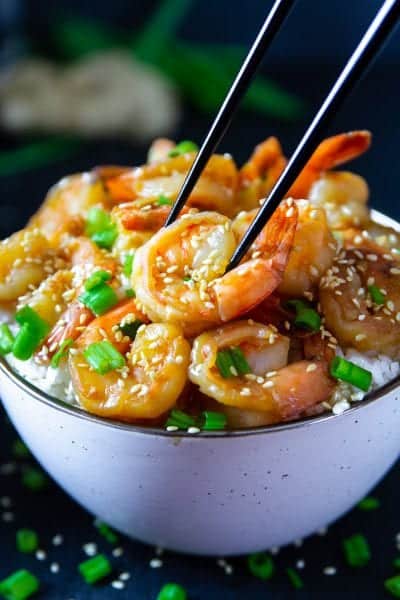 garlic ginger shrimp with green onion and sesame seeds on top in a white bowl with black chop sticks picking up the shrimp