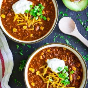 easy beef chili in 2 white bowls with sour cream, shredded cheddar, chopped green onion, and a sliced avocado on the side
