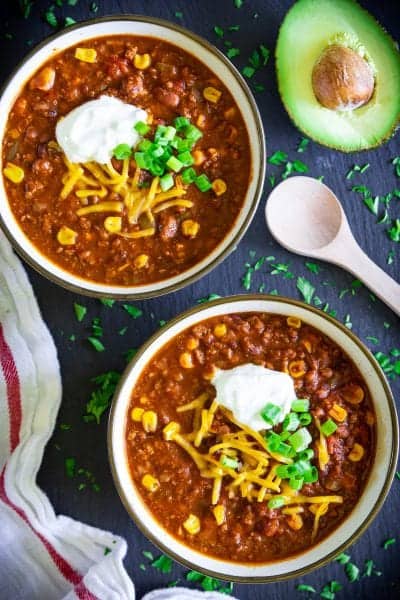easy beef chili in 2 white bowls with sour cream, shredded cheddar, chopped green onion, and a sliced avocado on the side