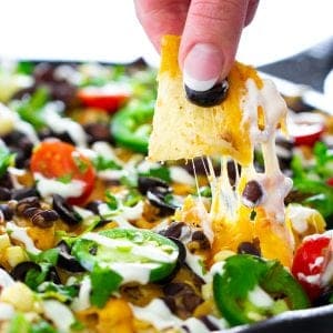 a close up shot of chicken nachos with a tortilla chip and cheddar cheese being pulled out of the skillet