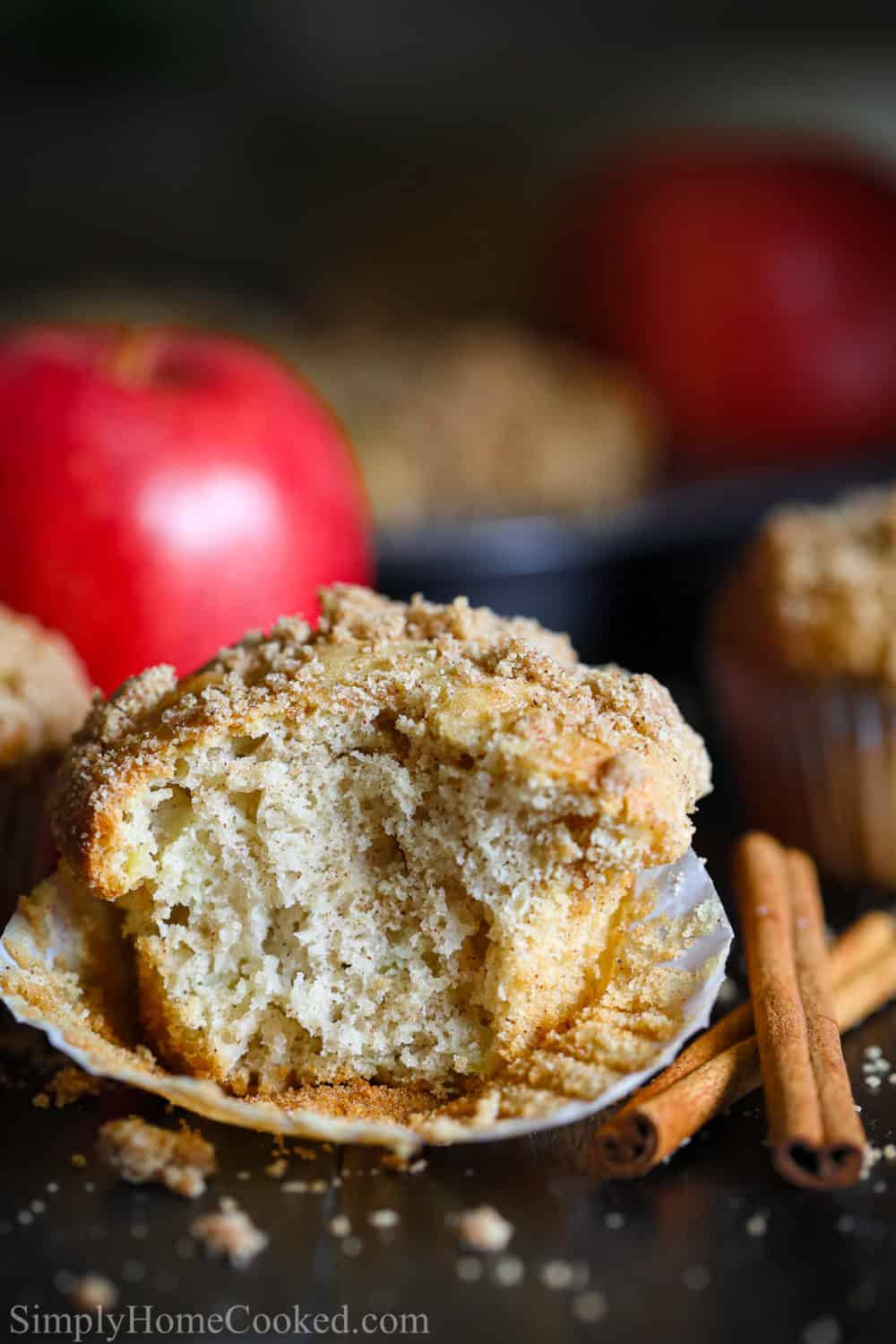 A single apple muffin that is partially bitten. An apple behind the muffin and 2 cinnamon sticks next to the muffin