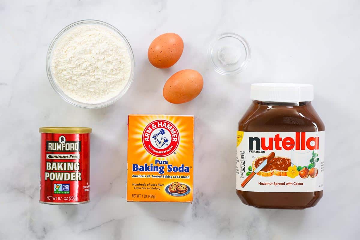 Soft and chewy Nutella cookies that are super easy to make and have an intense chocolate Nutella flavor. These are always a crowd pleaser. Which makes them great for parties! #nutella #nutellacookies #chocolatecookies #chewynutellacookies #easycookies overhead image of ingredients for Nutella cookies on a white background