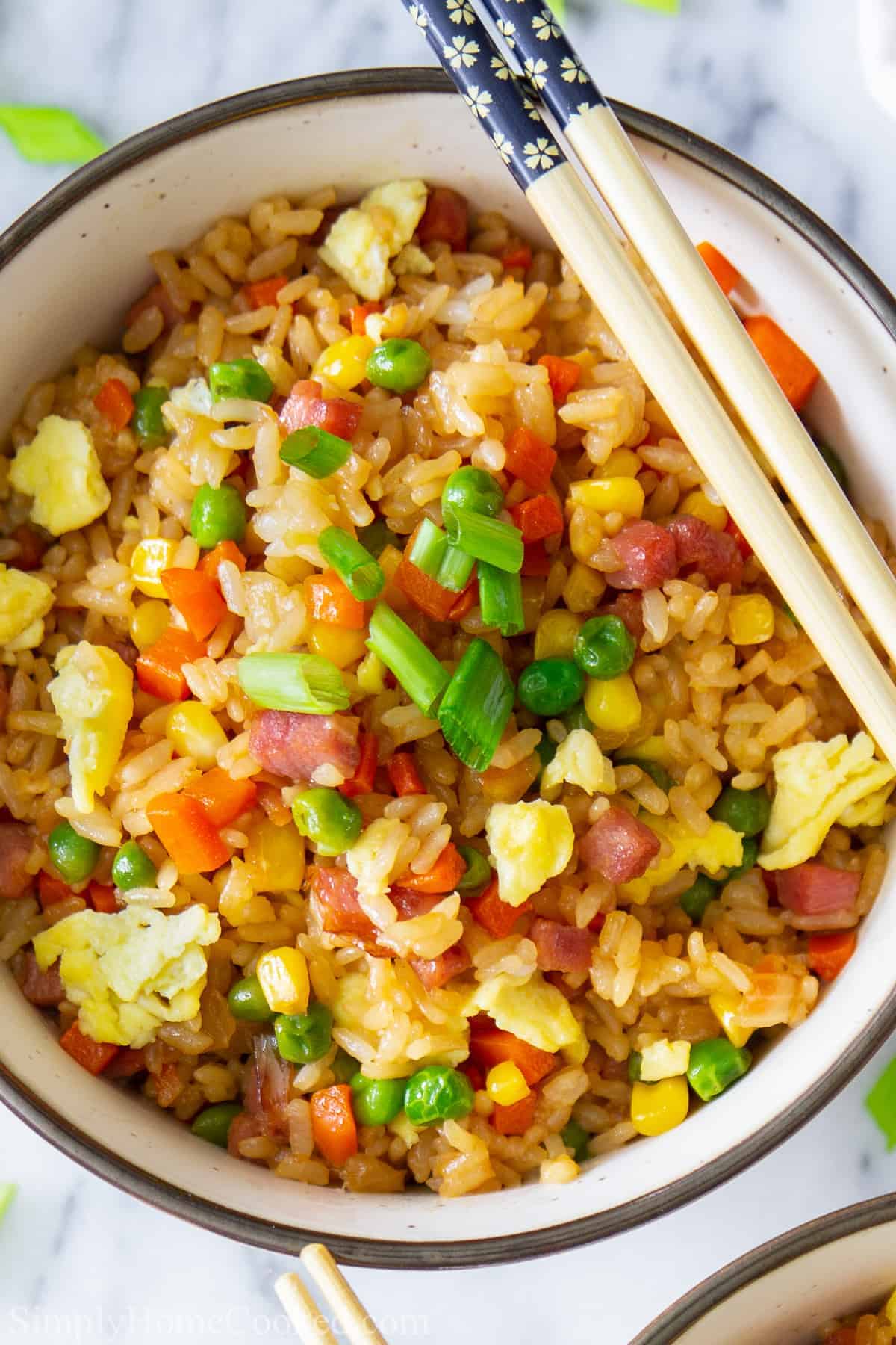 overhead image of pork fried rice in a bowl with green onion, peas, scrambled eggs, and corn on top. With chop sticks beside it.