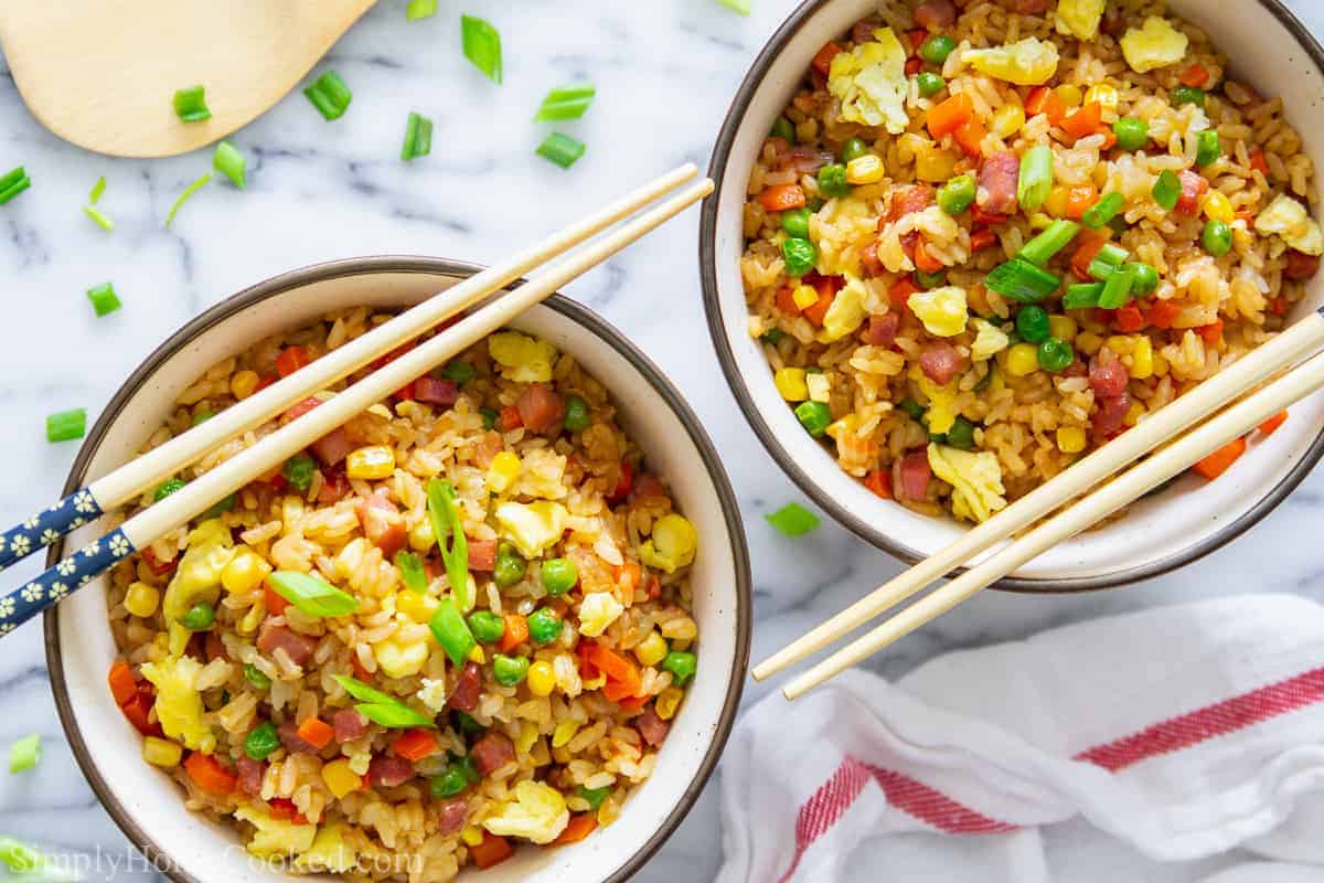 overhead image of pork fried rice in two bowls with green onion, peas, scrambled eggs, and corn on top. With chop sticks beside it.