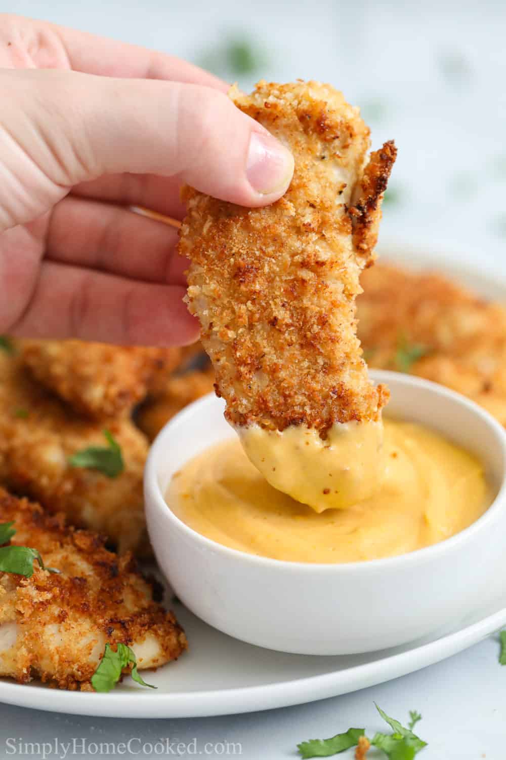 Breaded air fryer chicken tender being dipped into sauce with plate of chicken tenders in the background.