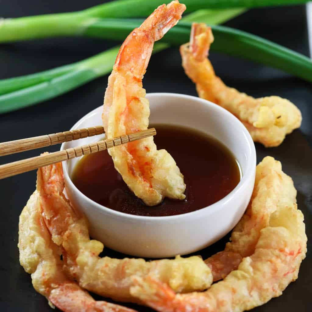 Shrimp tempura around a white bowl with tempura dipping sauce while one shrimp is dipped into it and there are green onions in the background.