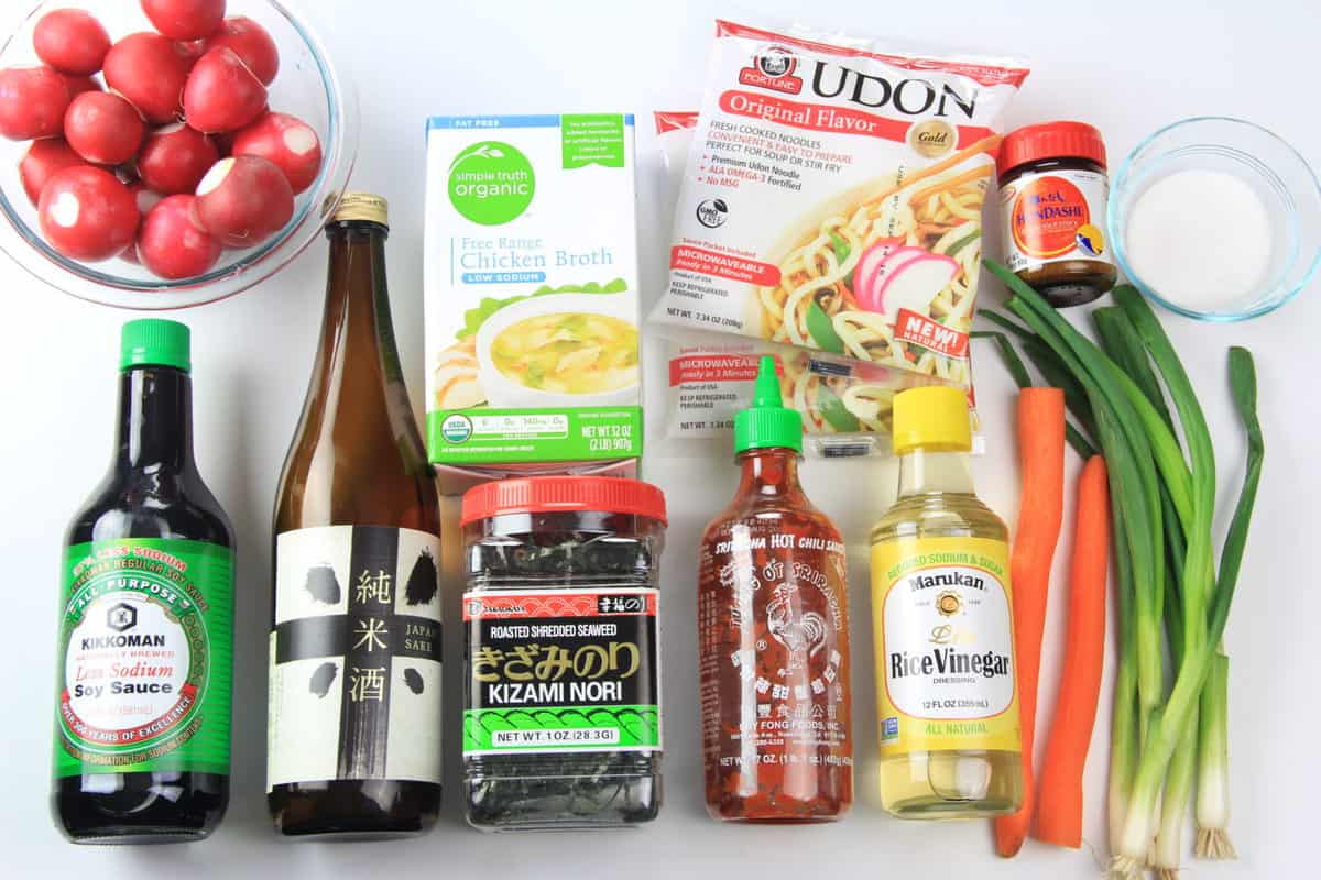 Ingredients to make udon soup, including soy sauce, sake, rice wine vinegar, Sriracha, radishes, udon noodles, carrots, green onion, hon-dashi powder, chicken stock, and seasoning.