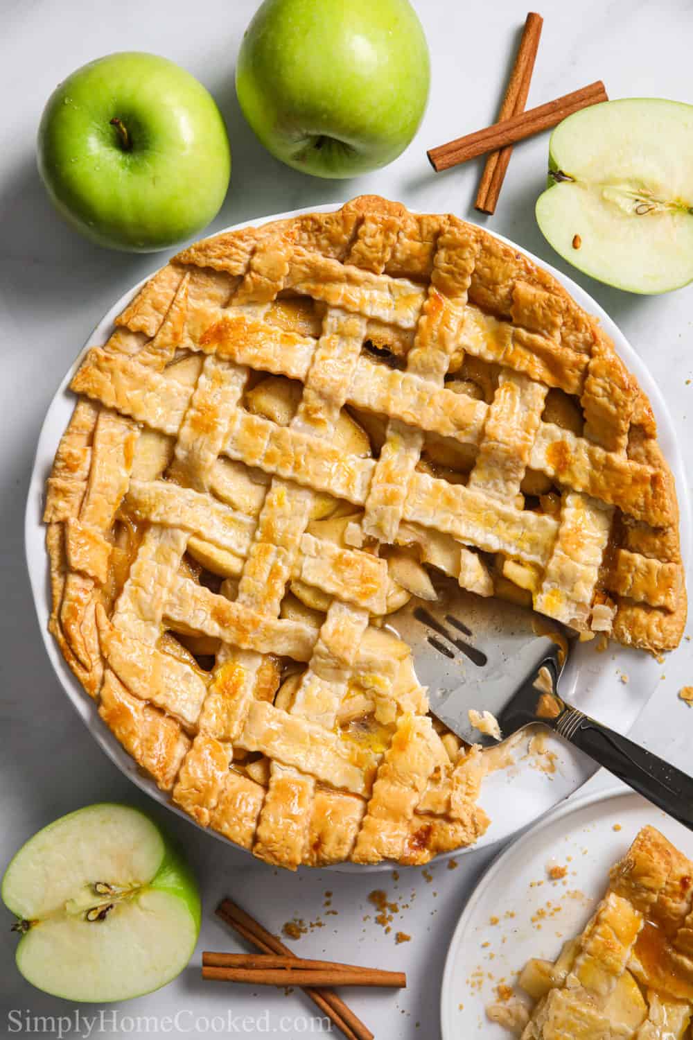 Homemade apple pie with a slice cut out and some green apples and cinnamon sticks nearby.